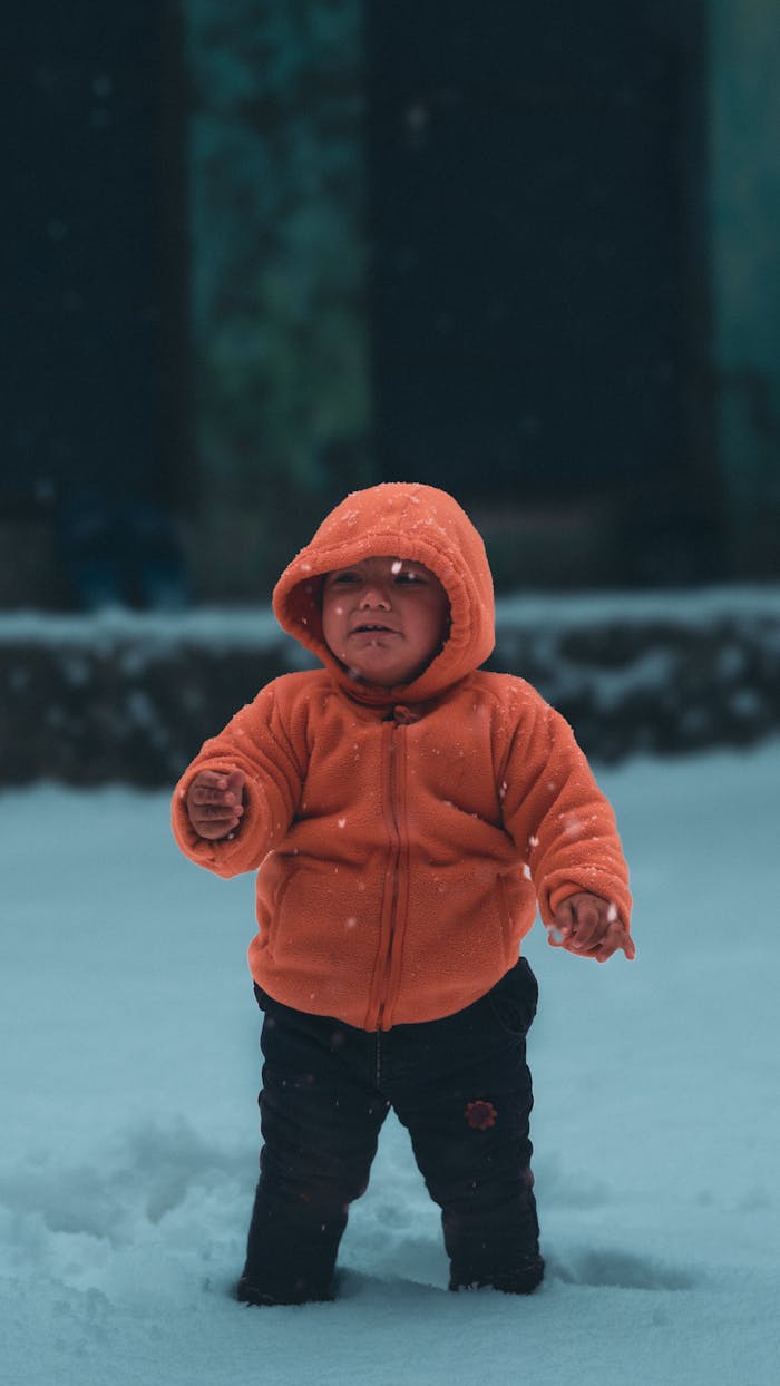 A young child in a bright orange jacket stands in the snow during a winter day outdoors.